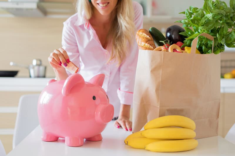 Woman with blond hair puts money in her piggy bank with a bag of groceries on the kitchen counter to