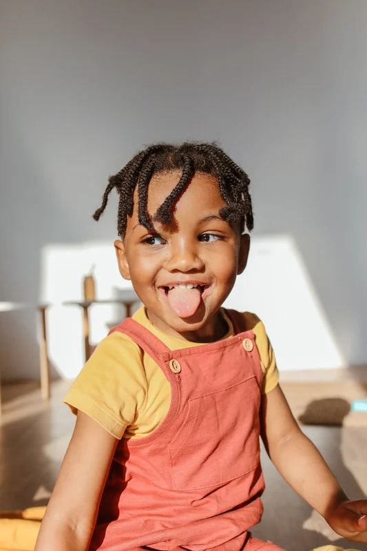 A cheeky toddler with braids and orange dungarees sticking his tongue out 