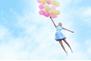 Woman in a blue dress floating up to the sky clasping a big bunch of pastel-coloured balloons