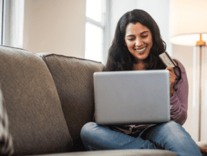 A brown-haired woman sits on her couch at home. She has a laptop propped on her knees while she works on her savings plan.