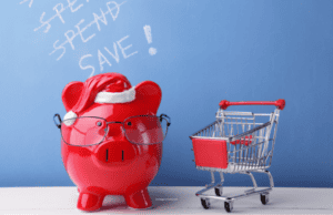 A red piggy bank wearing a Santa hat and a pair of glasses sands next to a shopping cart. a banner behind reads 'Save!'